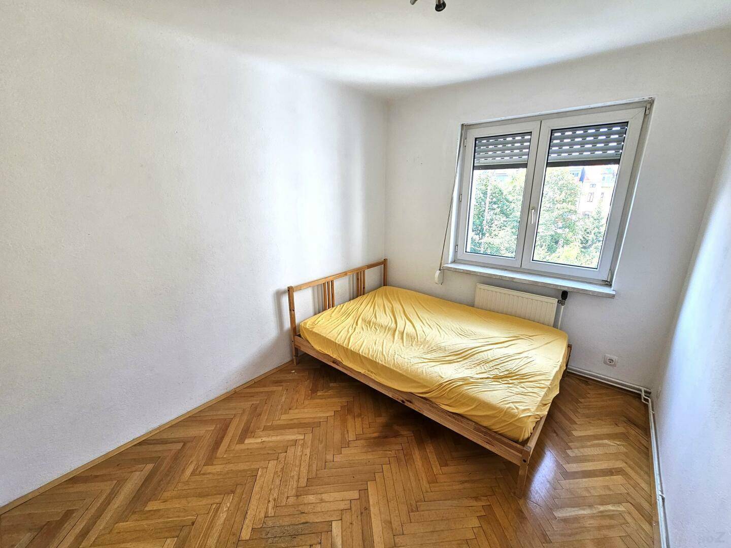 Schlafzimmer mit Fischgrätparkett, Einzelbett und Fenster mit Blick ins Grüne.