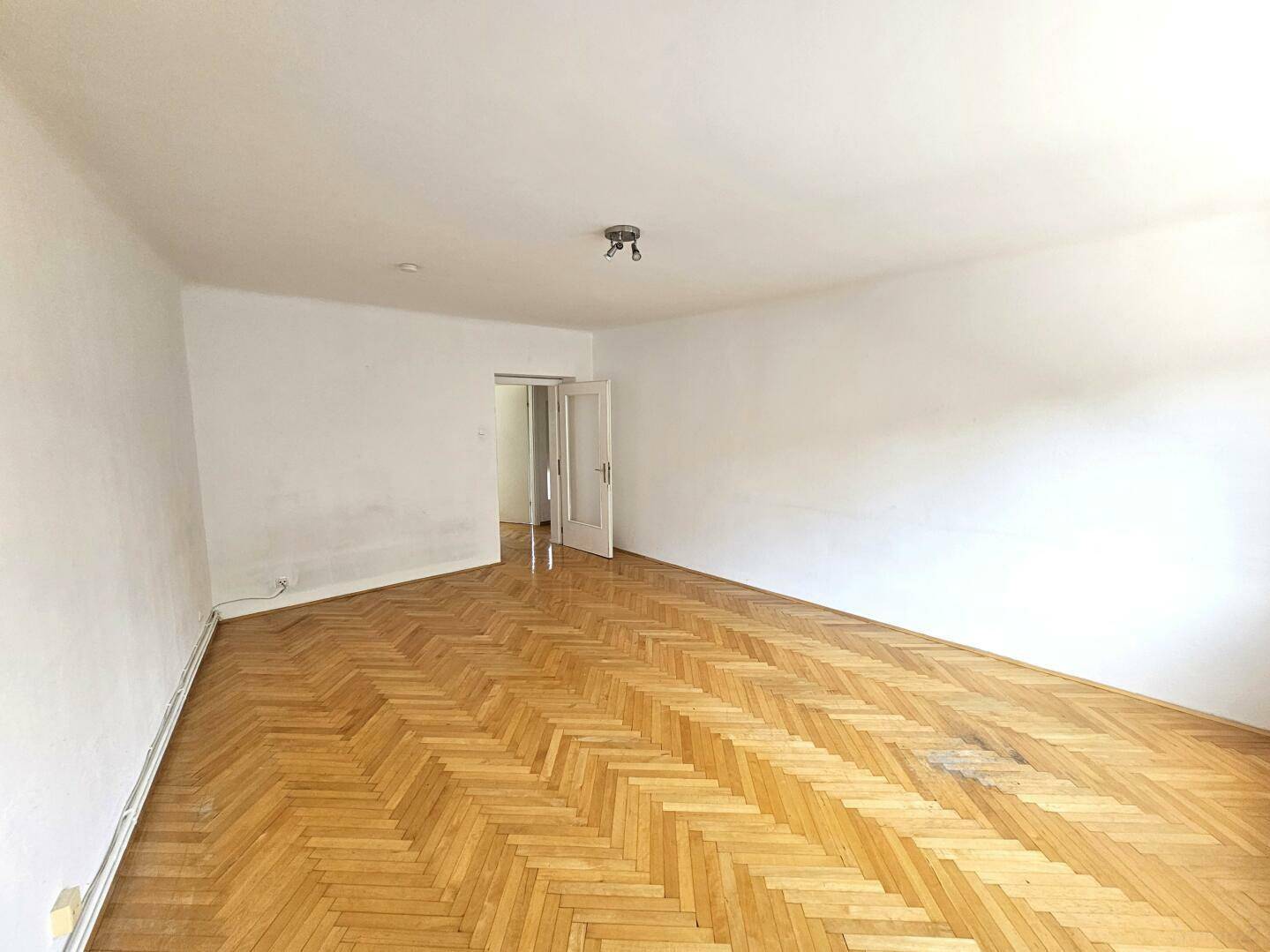 Geräumiges Wohnzimmer mit Fischgrätparkett und Zugang zu einem weiteren Raum.