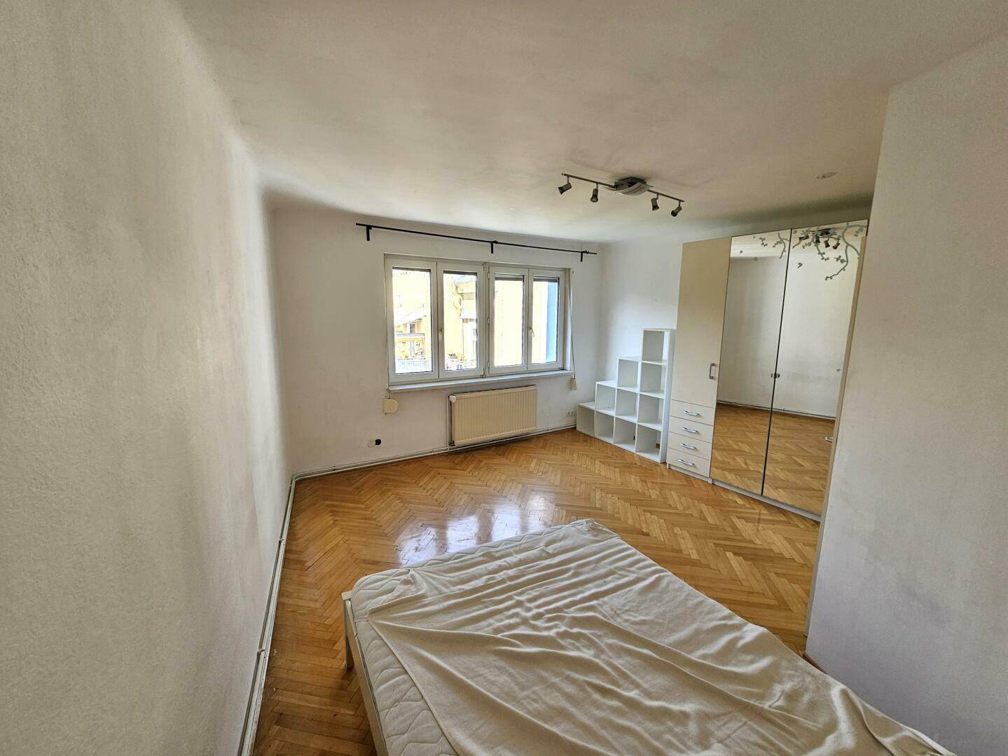 Schlafzimmer mit Parkettboden, Fenster und einem weißen Bettgestell.