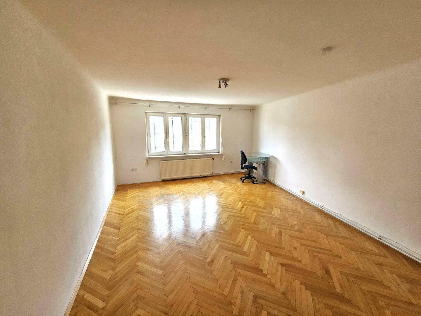 Helles Wohnzimmer mit Fischgrätparkettboden und einem kleinen Arbeitsbereich am Fenster.