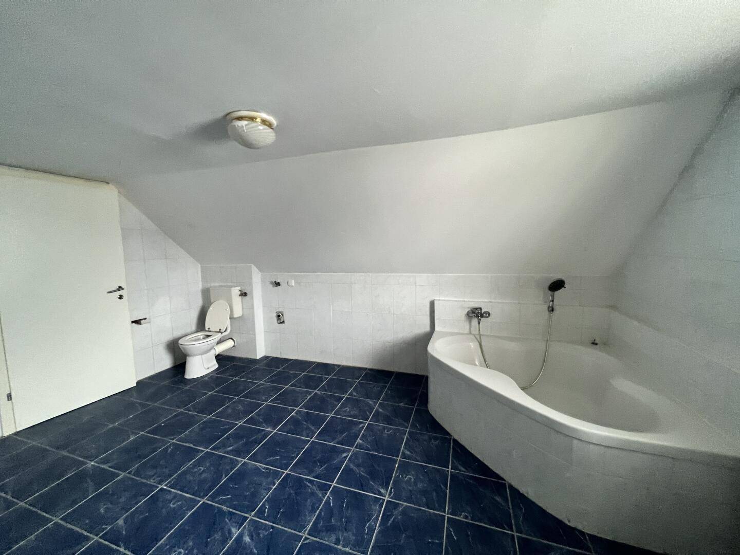 Helles Badezimmer im Dachgeschoss mit blauen Bodenfliesen, Toilette und großer Eckbadewanne.