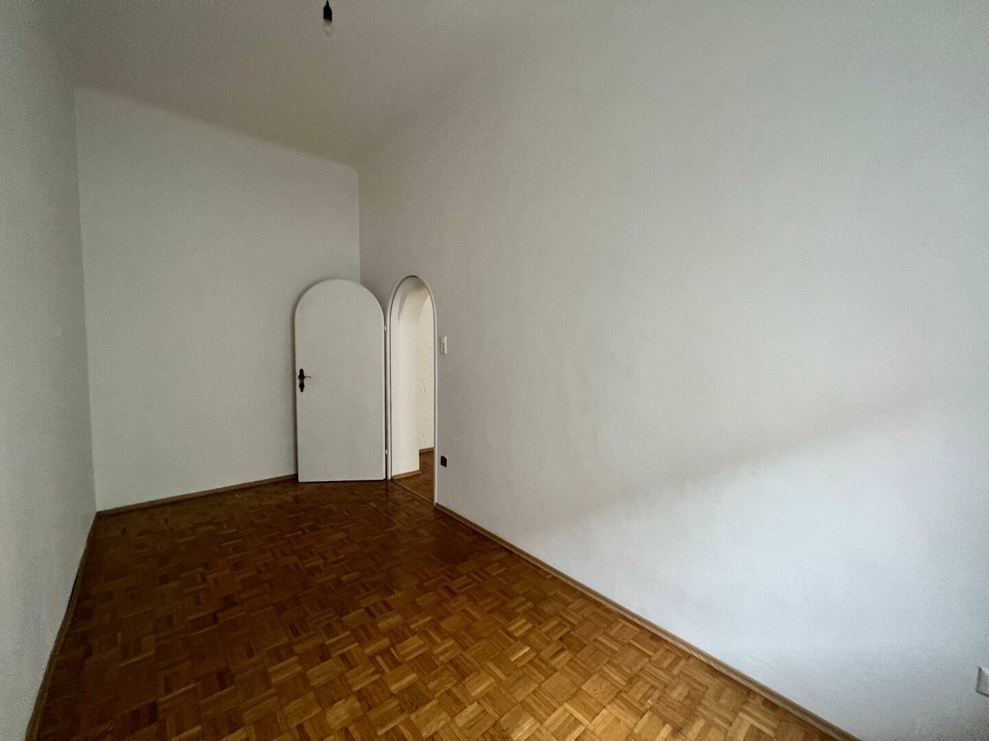 Leeres Zimmer mit Fischgrätparkett und zwei Rundbögen, die zu weiteren Räumen führen.