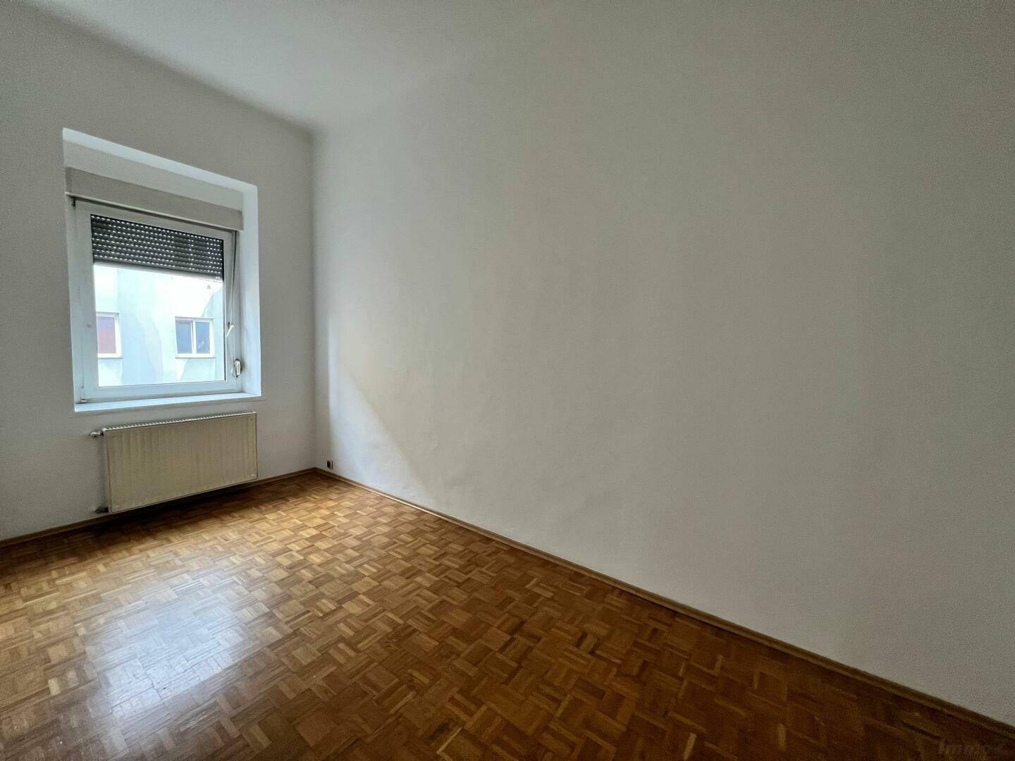 Leeres Zimmer mit klassischem Parkettboden und einem Fenster mit Rollladen.