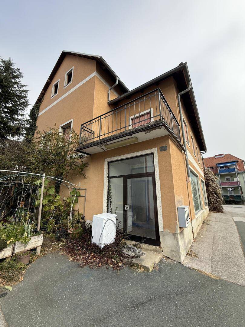Gebäudeaußenansicht mit Balkon und Erdgeschosszugang, umgeben von Vegetation und einer Straße.