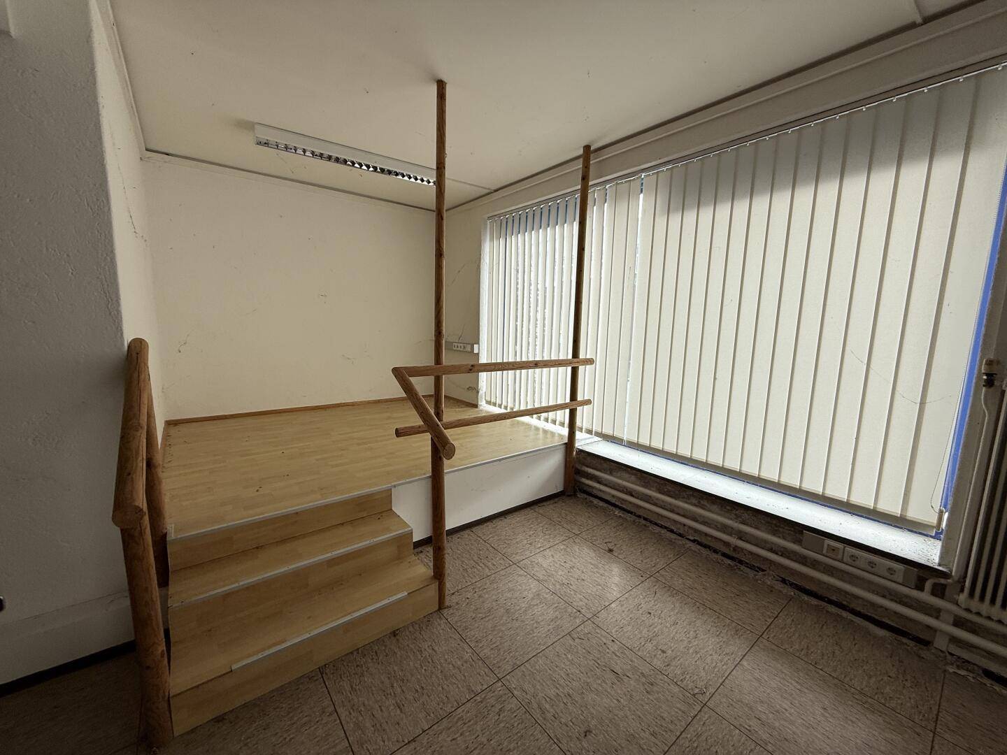 Innenraum mit erhöhtem Holzpodest, Treppe und Fenster mit Jalousien, auf einem gefliesten Boden.