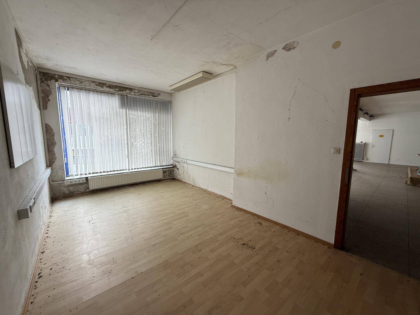 Kleiner Innenraum mit Holzfußboden, Fenster mit Jalousien und beschädigten Wänden, renovierungsbedürftig.
