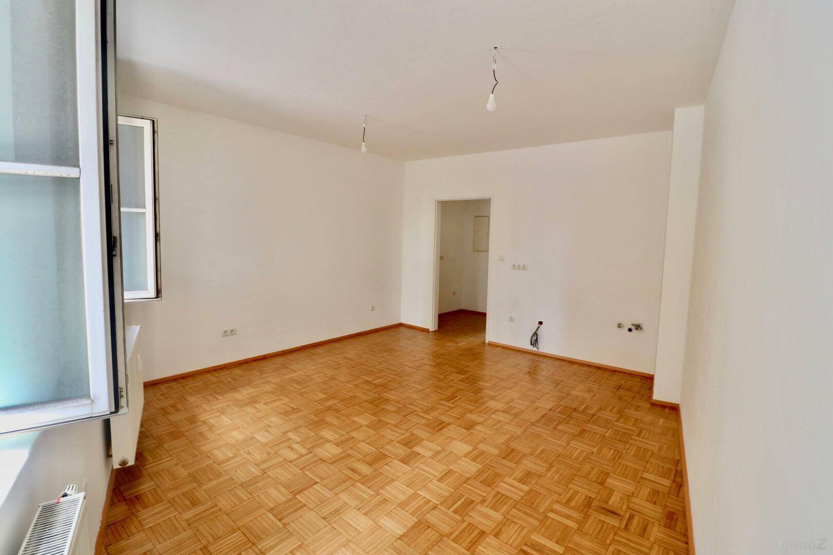 Geräumiges, leeres Zimmer mit Parkettboden und viel Tageslicht durch die Fenster.