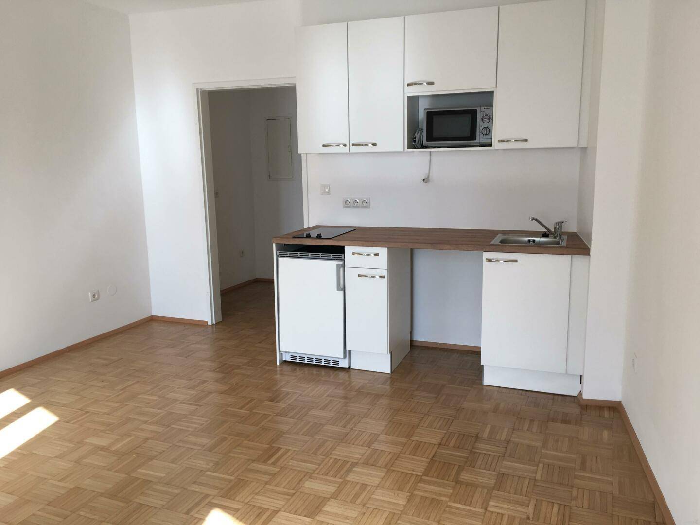 Kompakte Küchenzeile mit weißen Schränken, Mikrowelle und Kochfeld in einem Wohnraum.