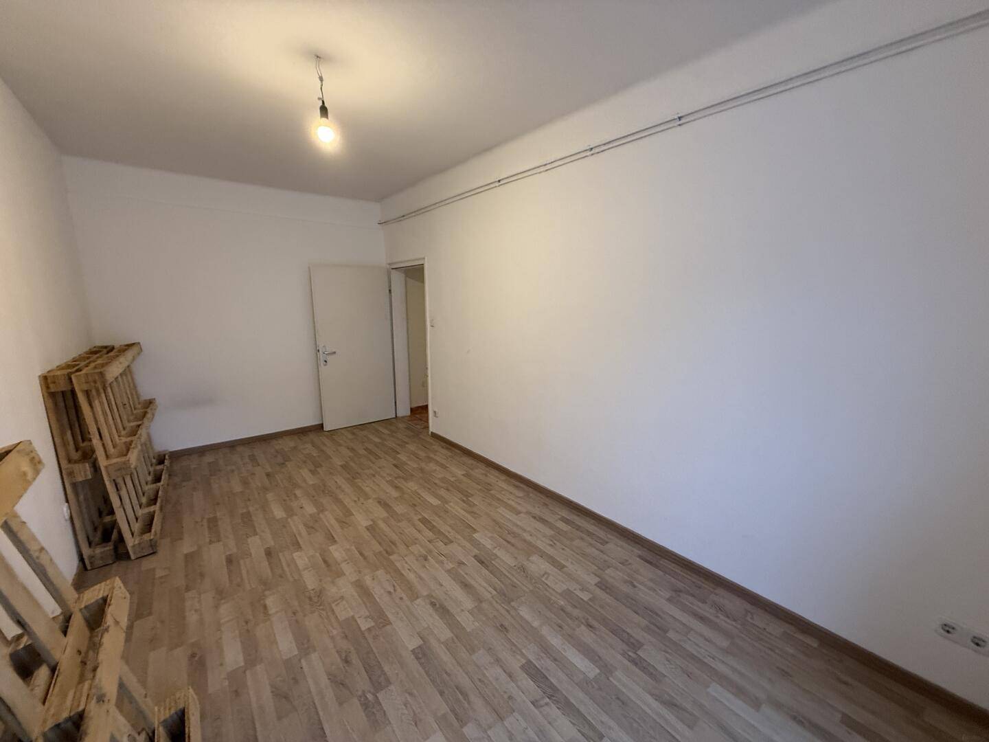 Leerer Raum mit Laminatboden, weißen Wänden und einfacher Deckenleuchte.