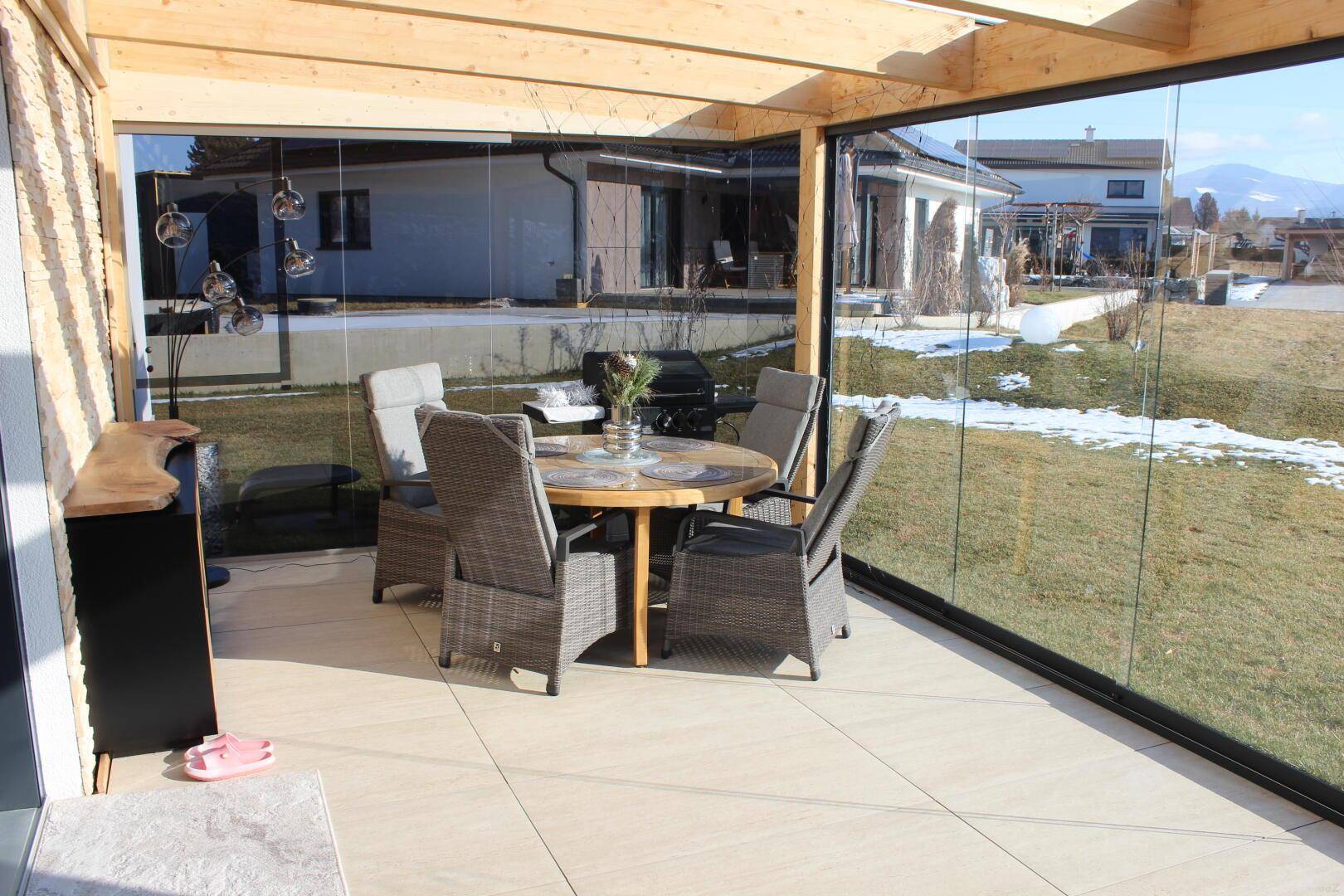 Heller Wintergarten mit Esstisch und Stühlen, umgeben von Glaswänden mit Blick ins Grüne.