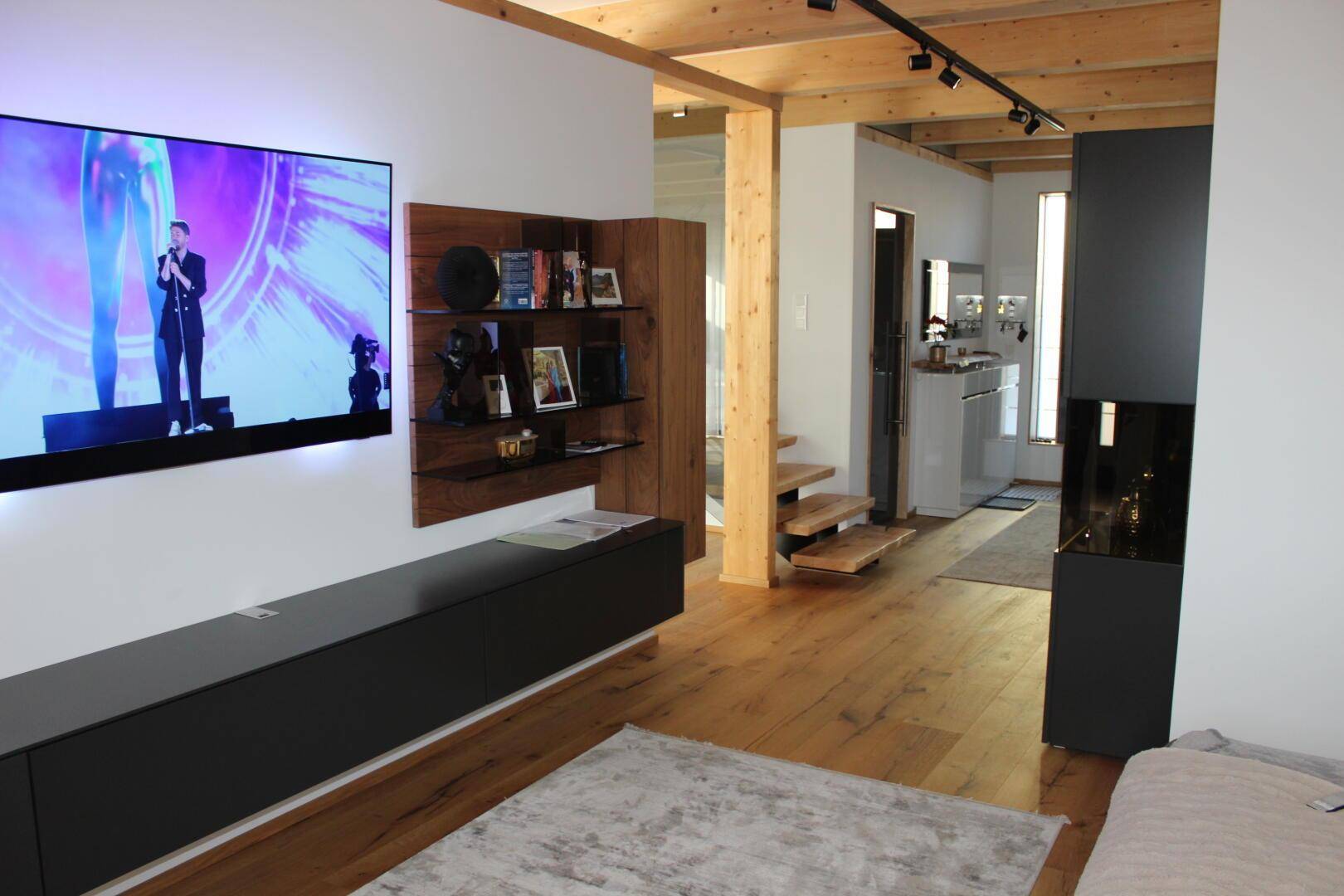 Offener Wohnbereich mit Holzboden, Wandfernseher und Blick zum Eingangsbereich und Treppe.