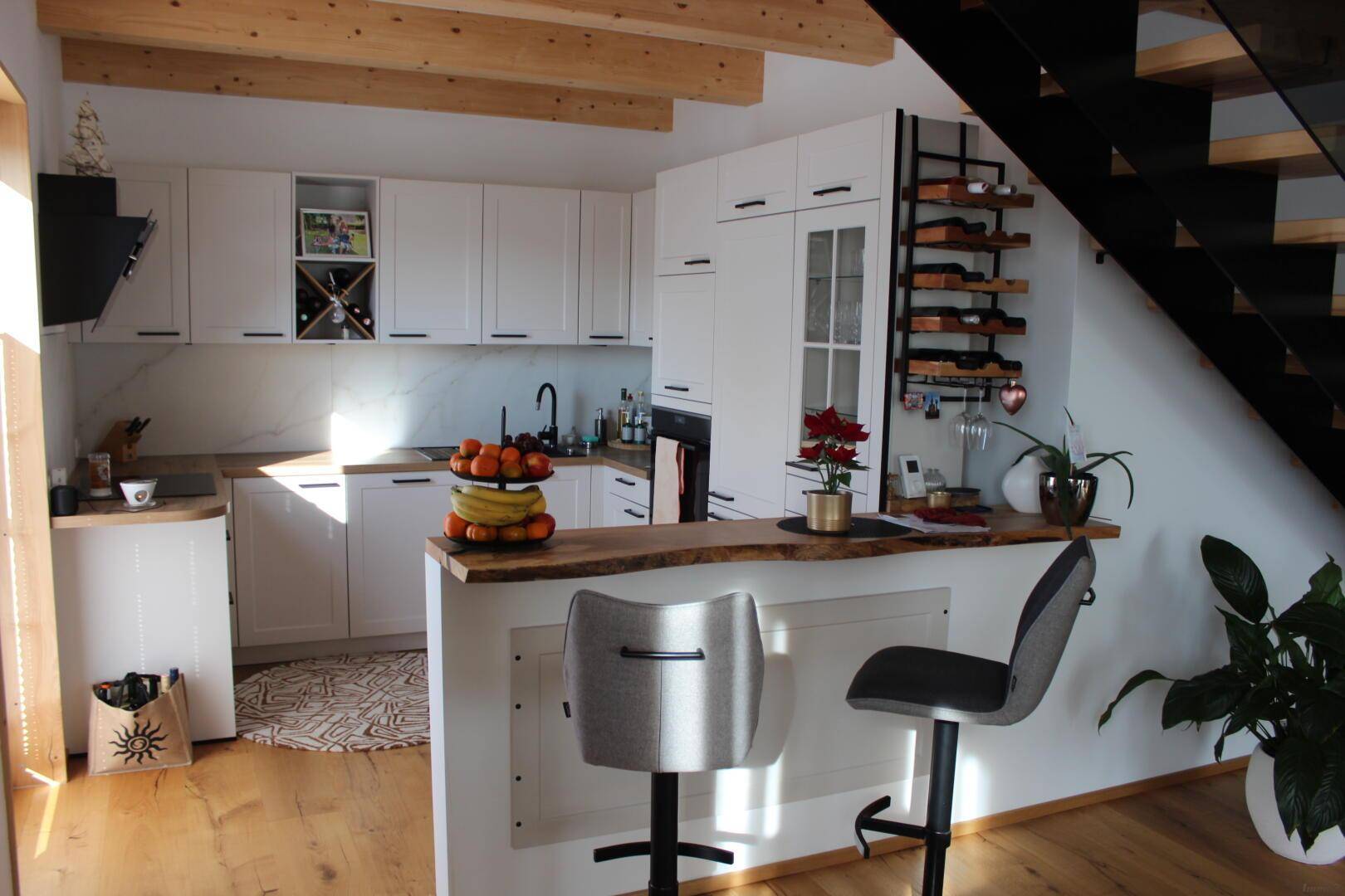 Moderne weiße Küche mit Holzarbeitsplatte, Barhockern und integriertem Weinregal unter der Treppe.