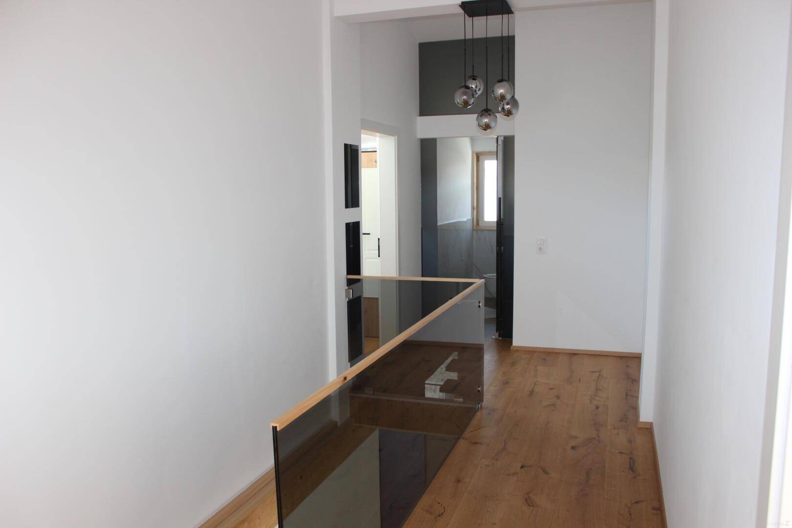 Heller Flur im Obergeschoss mit Glasgeländer, Holzboden und moderner Hängelampe.