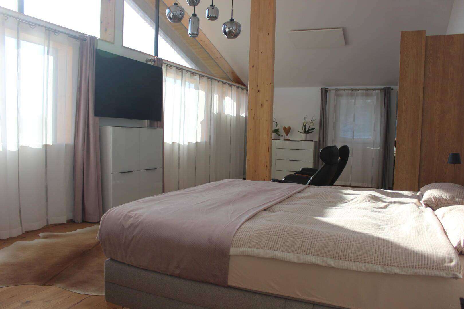 Helles Schlafzimmer mit Doppelbett, großen Fenstern und sichtbaren Holzbalken unter dem Dach.