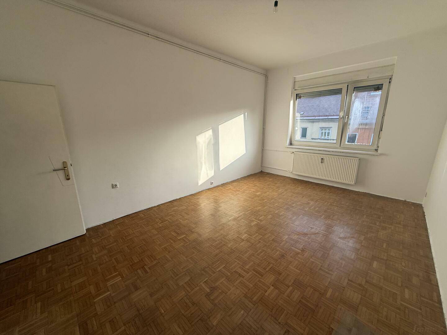 Helles Zimmer mit klassischem Parkettboden und Fenster, das viel Sonnenlicht hereinlässt.