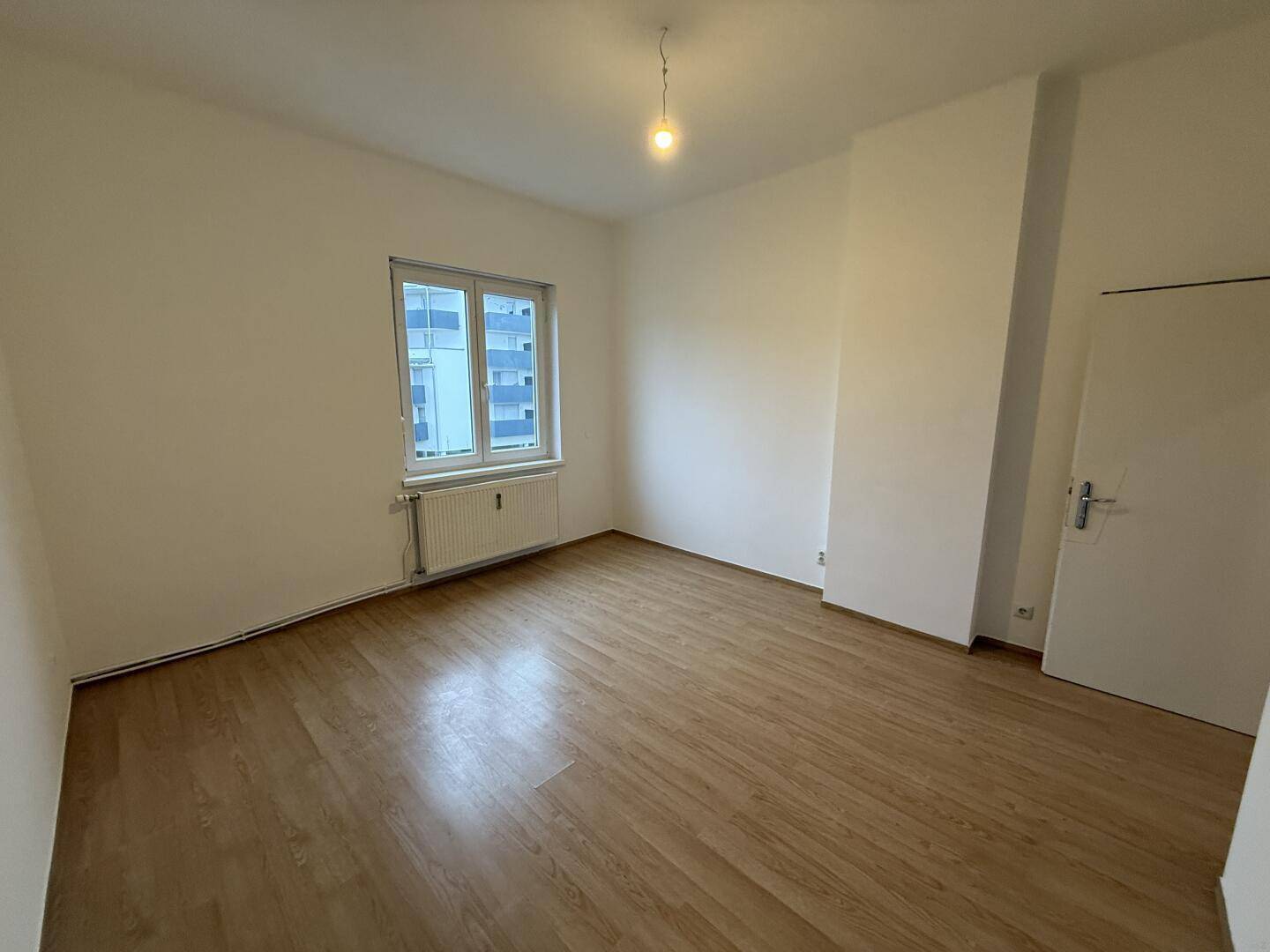Geräumiges, helles Zimmer mit Laminatboden und Blick auf umliegende Gebäude.