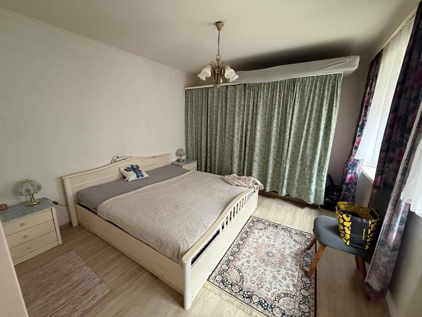 Gemütliches Schlafzimmer mit hellem Bett, Nachttischen, Teppich und Vorhängen am Fenster.
