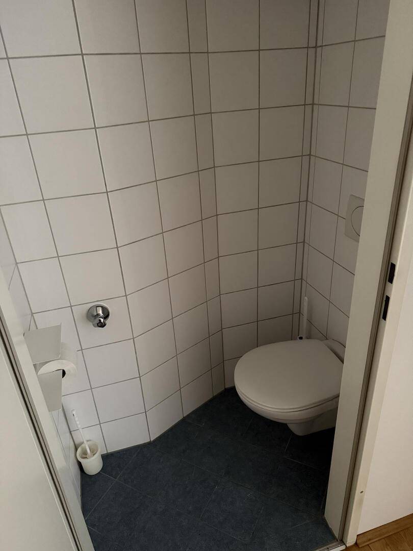 Kleine separate Toilette mit weißen Wandfliesen und dunklem Boden, ausgestattet mit einer Wandtoilette.