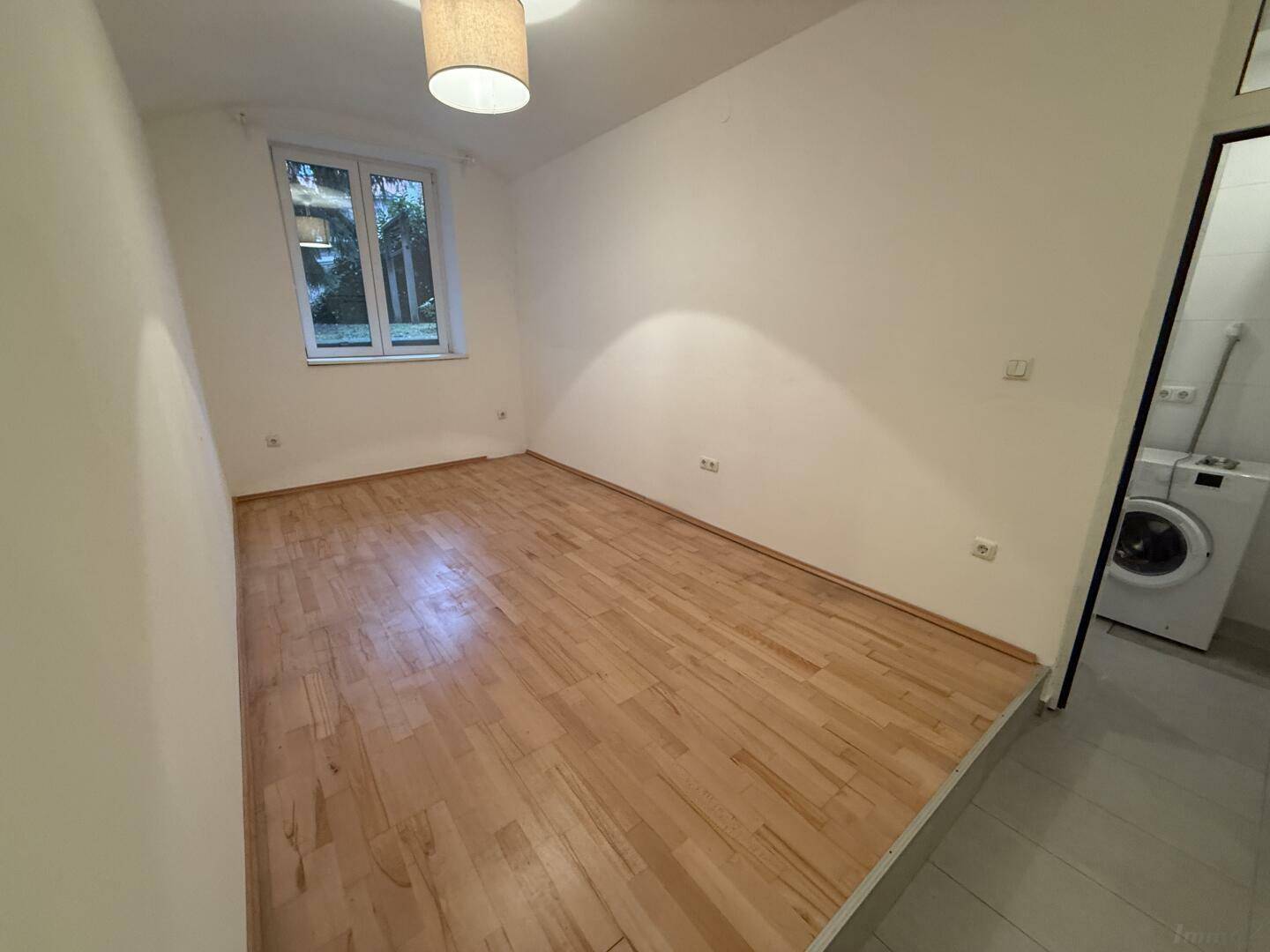 Helles Zimmer mit Parkettboden und Fenster, angrenzend an einen Bereich mit Waschmaschine.