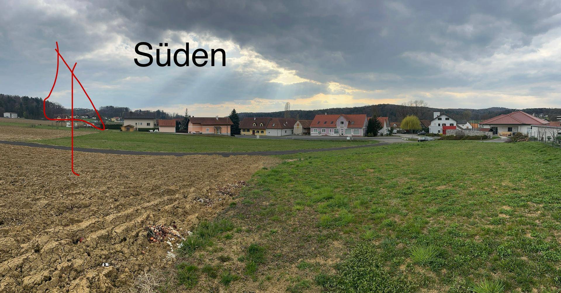 Panoramablick nach Süden über ein Ackerfeld mit Häusern und bewaldeten Hügeln in der Ferne.