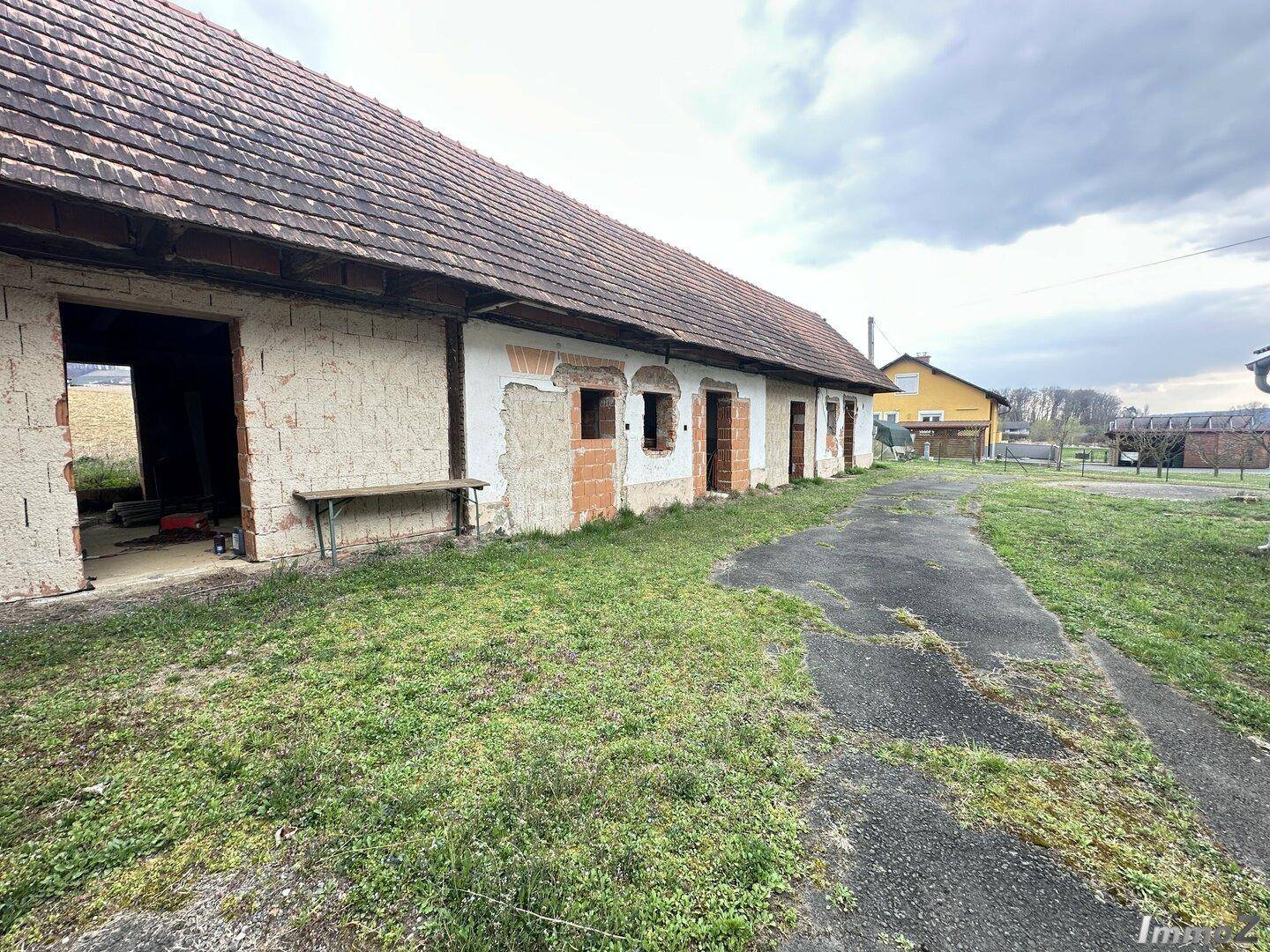 Lange, baufällige Scheune mit Ziegeldach und sichtbaren Mauerwerksschäden in ländlicher Umgebung unter bewölktem Himmel.