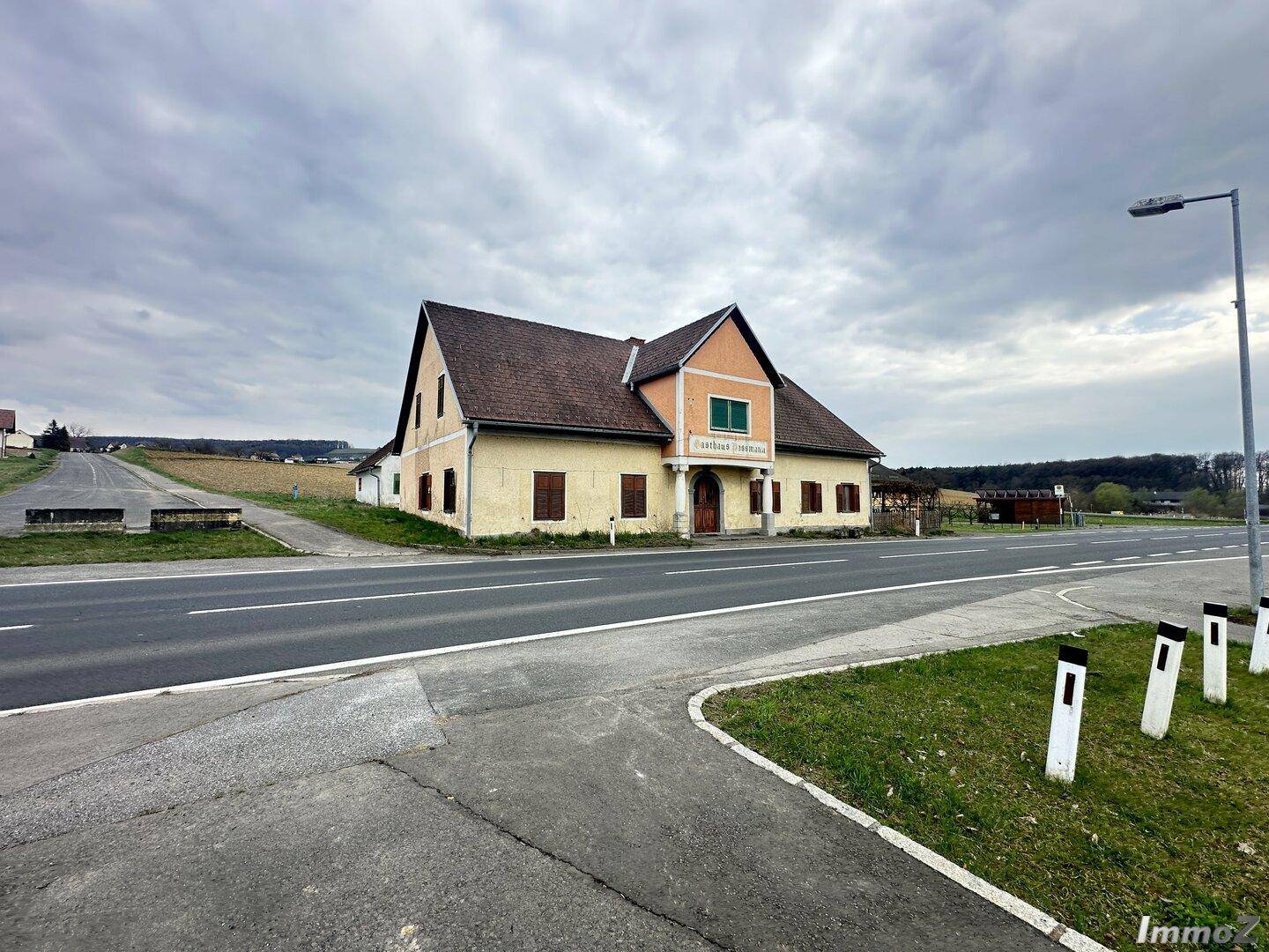 Traditionelles Gebäude mit Satteldach und Fensterläden an einer Landstraße unter bewölktem Himmel.