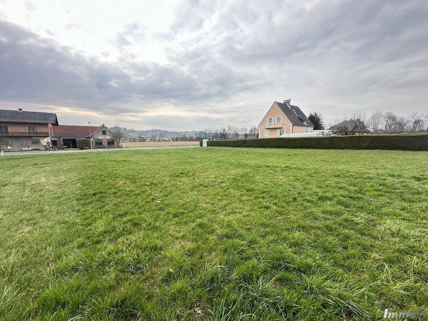 Weitläufige grüne Wiesen und Felder mit vereinzelten Häusern in der Ferne unter bewölktem Himmel.