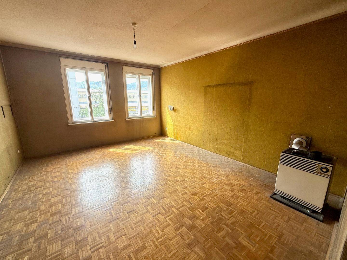 Helles Wohnzimmer mit Parkettboden, gelben Wänden und einem Heizofen, bereit zur Modernisierung.
