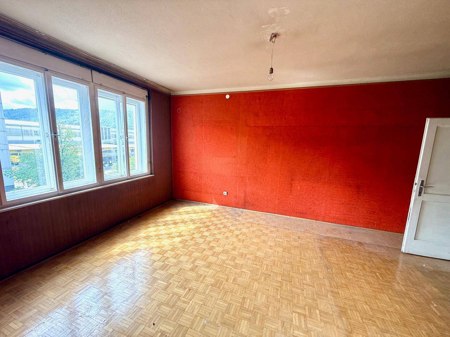 Wohnzimmer mit Parkettboden, roten Wänden und großen Fenstern, wartet auf Renovierung.
