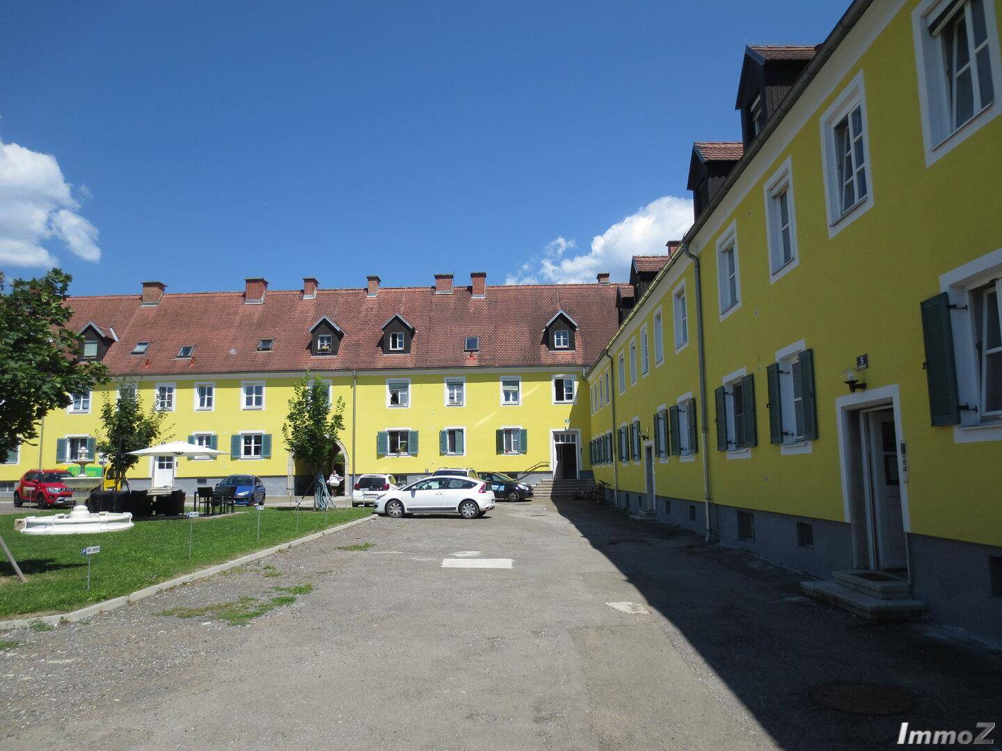 Gelbe Gebäude mit roten Dächern und grünen Fensterläden umgeben einen Innenhof mit Parkplätzen und einem Brunnen.