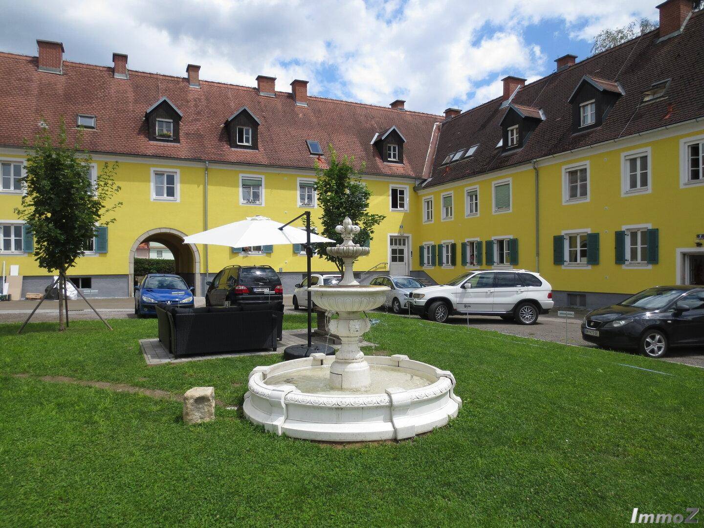 Innenhof mit einem dekorativen Brunnen, Rasenflächen und geparkten Autos vor den gelben Gebäuden.