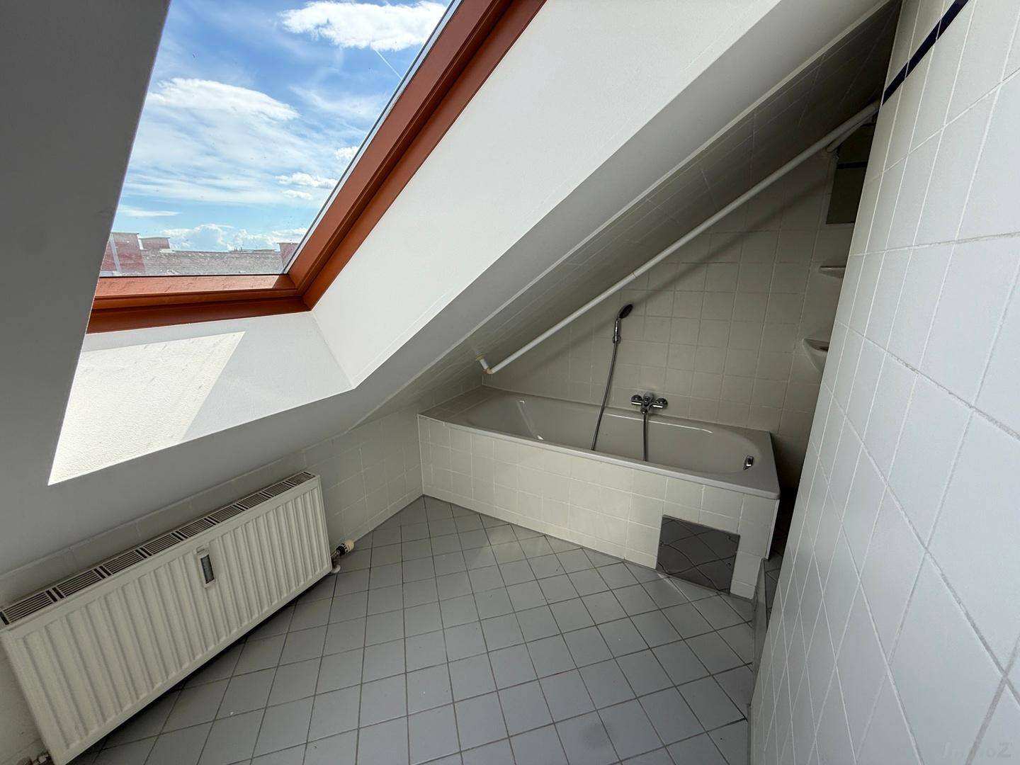 Badewannenbereich mit Dachfenster und Heizkörper, umgeben von weißen Fliesen und Dachschrägen.