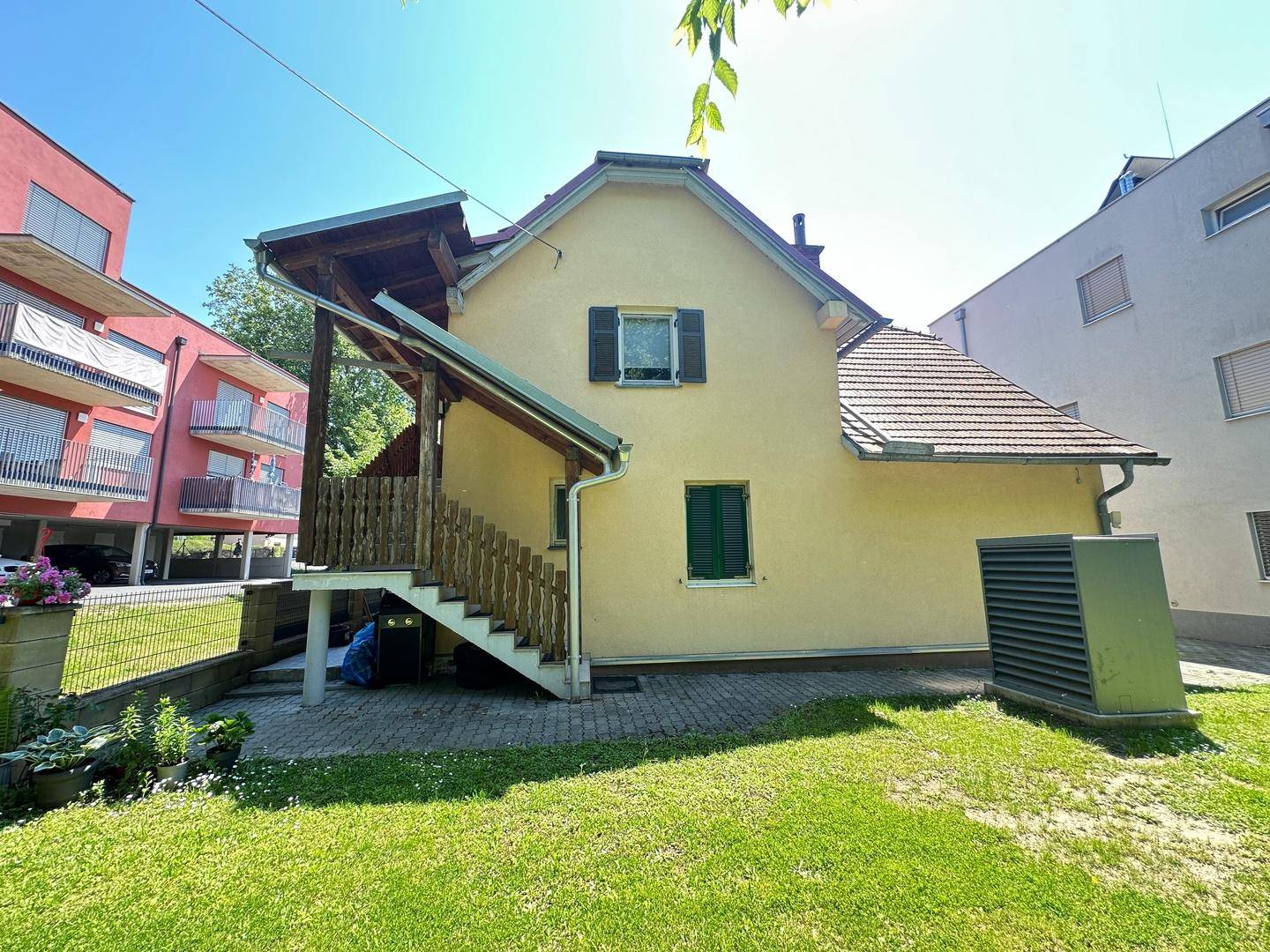 Rückansicht des traditionellen Hauses mit Außentreppe und einem Technikschrank im Gartenbereich.