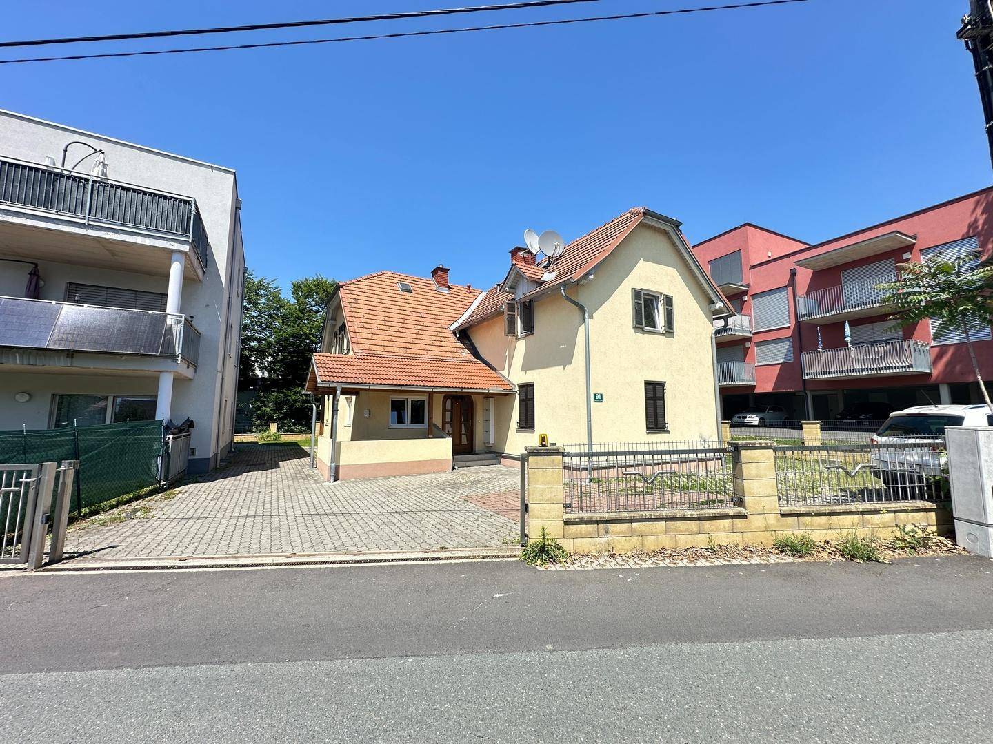 Traditionelles Haus mit Ziegeldach und gepflasterter Einfahrt, neben einem modernen Gebäude.
