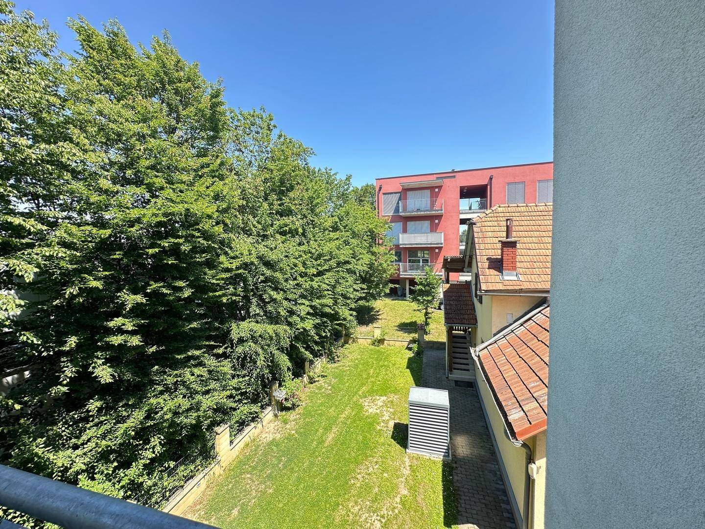 Blick vom Balkon auf den grünen Gartenbereich und das rote Nachbargebäude.