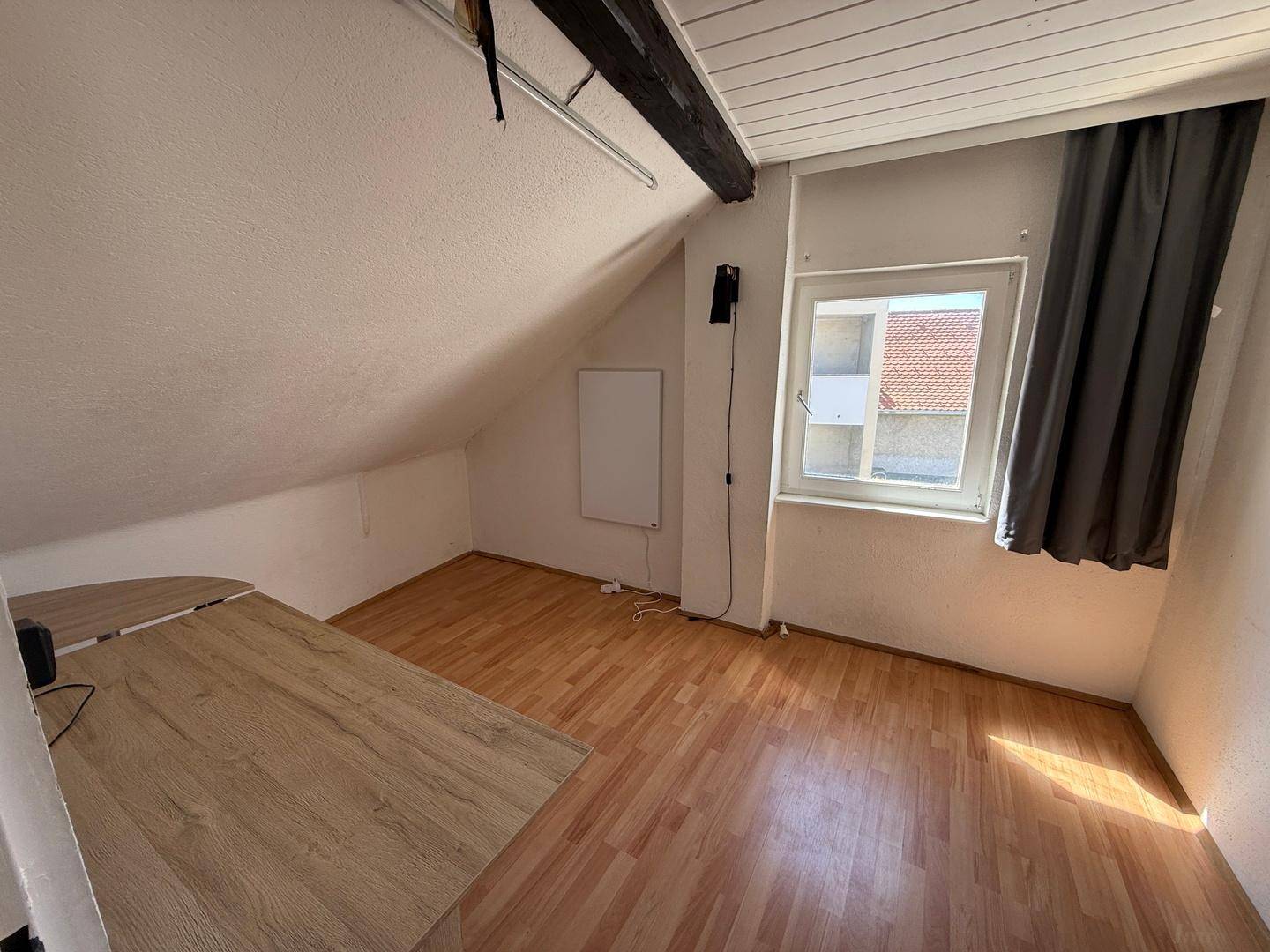 Helles Zimmer im Dachgeschoss mit Schreibtisch, Laminatboden und Fenster mit Blick nach draußen.