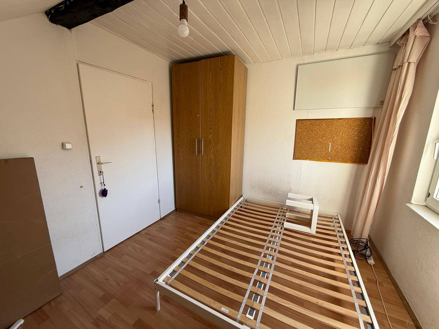 Schlafzimmer mit Bettgestell, Kleiderschrank und Fenster in einer Dachgeschosswohnung.