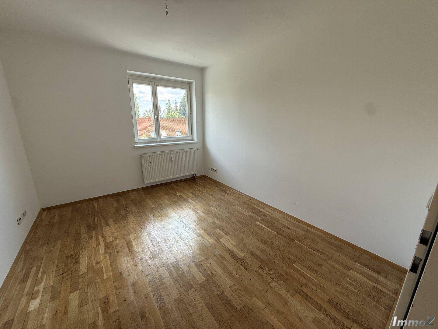 Leeres Schlafzimmer mit Holzboden und Fenster, das viel Tageslicht hereinlässt.