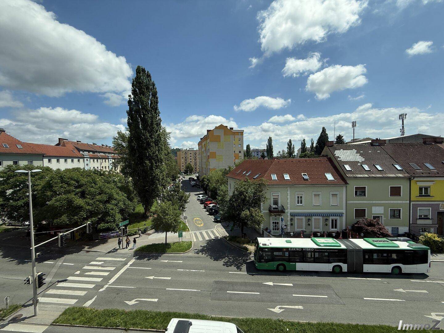 Blick auf eine belebte Straße mit Gebäuden, Bäumen und einem öffentlichen Bus unter blauem Himmel.