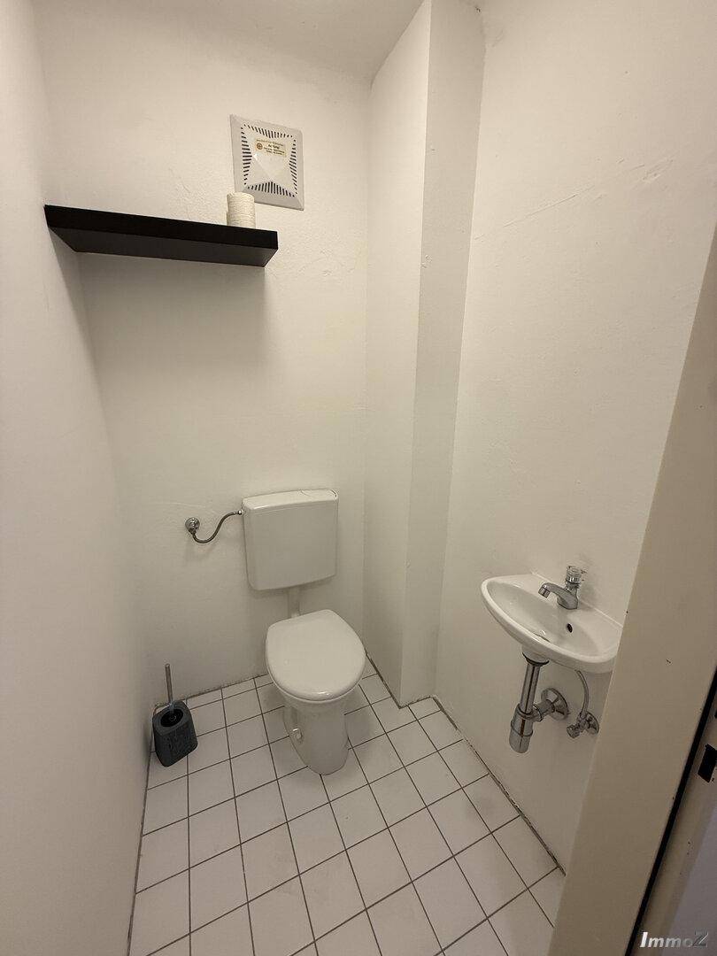 Separates WC mit Toilette, kleinem Waschbecken und weißen Fliesen an Boden und Wänden.