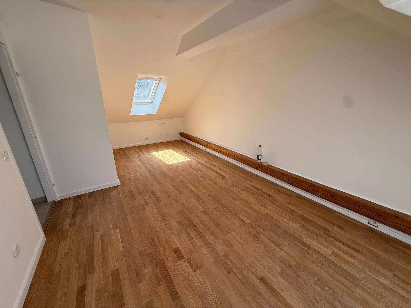 Helles Zimmer im Dachgeschoss mit Holzboden, schrägen Wänden und einem Dachfenster.