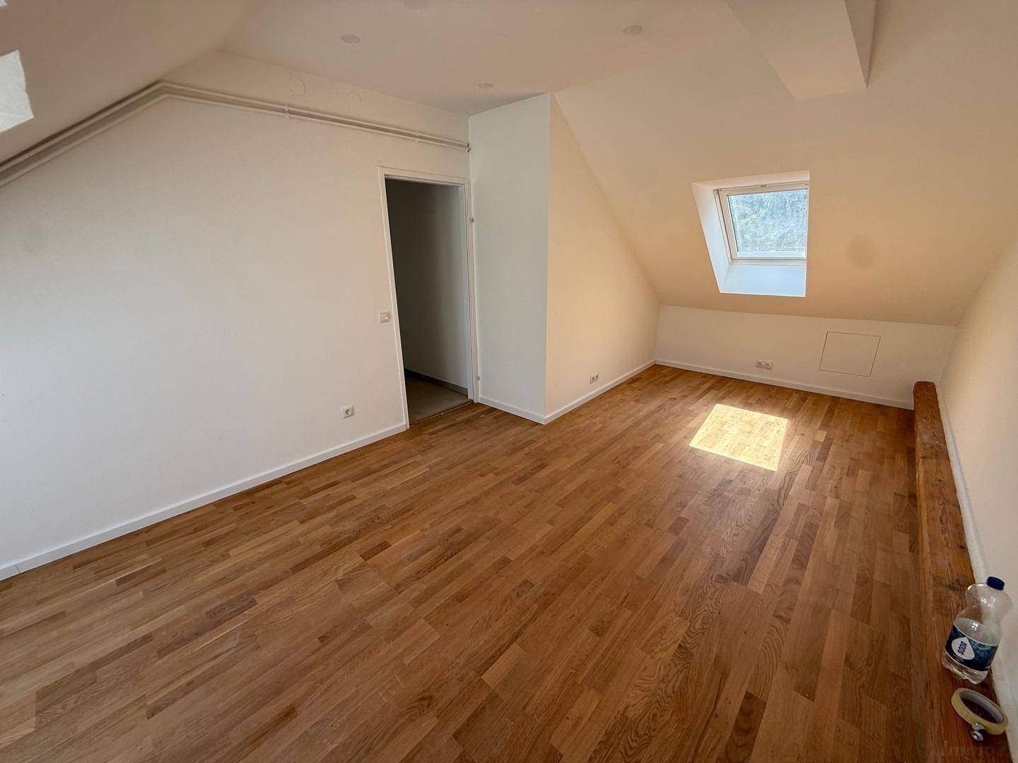 Geräumiges Dachzimmer mit Holzboden, weißen Wänden und einem Dachfenster, das den Raum erhellt.