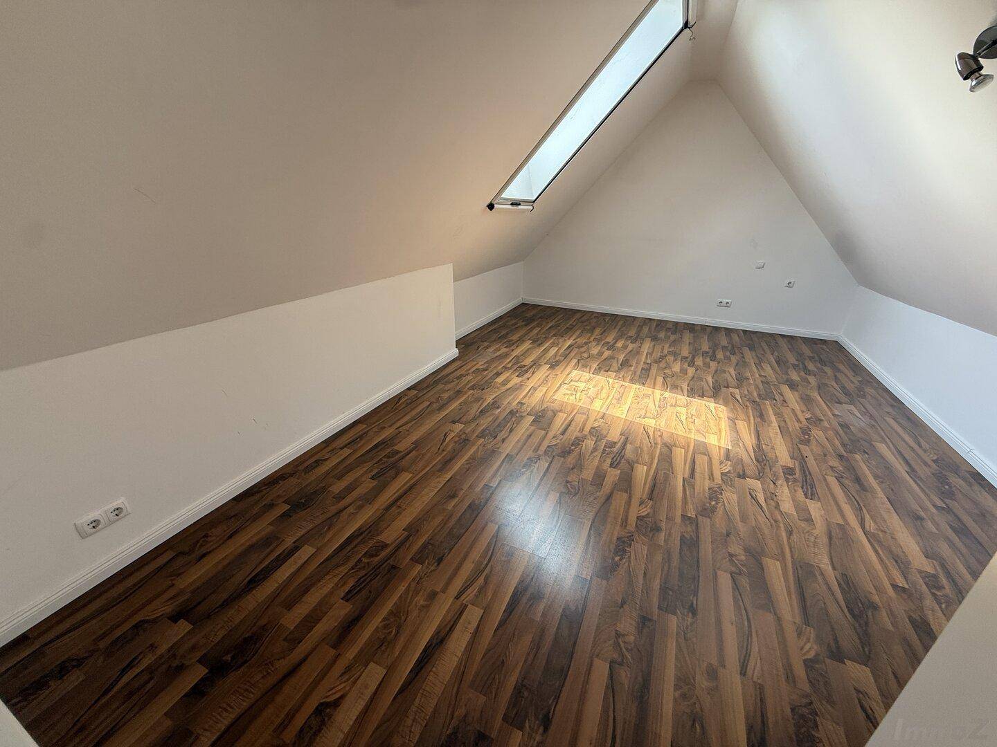 Schlafzimmer mit Laminatboden und Dachschräge, beleuchtet durch ein langes Dachfenster.