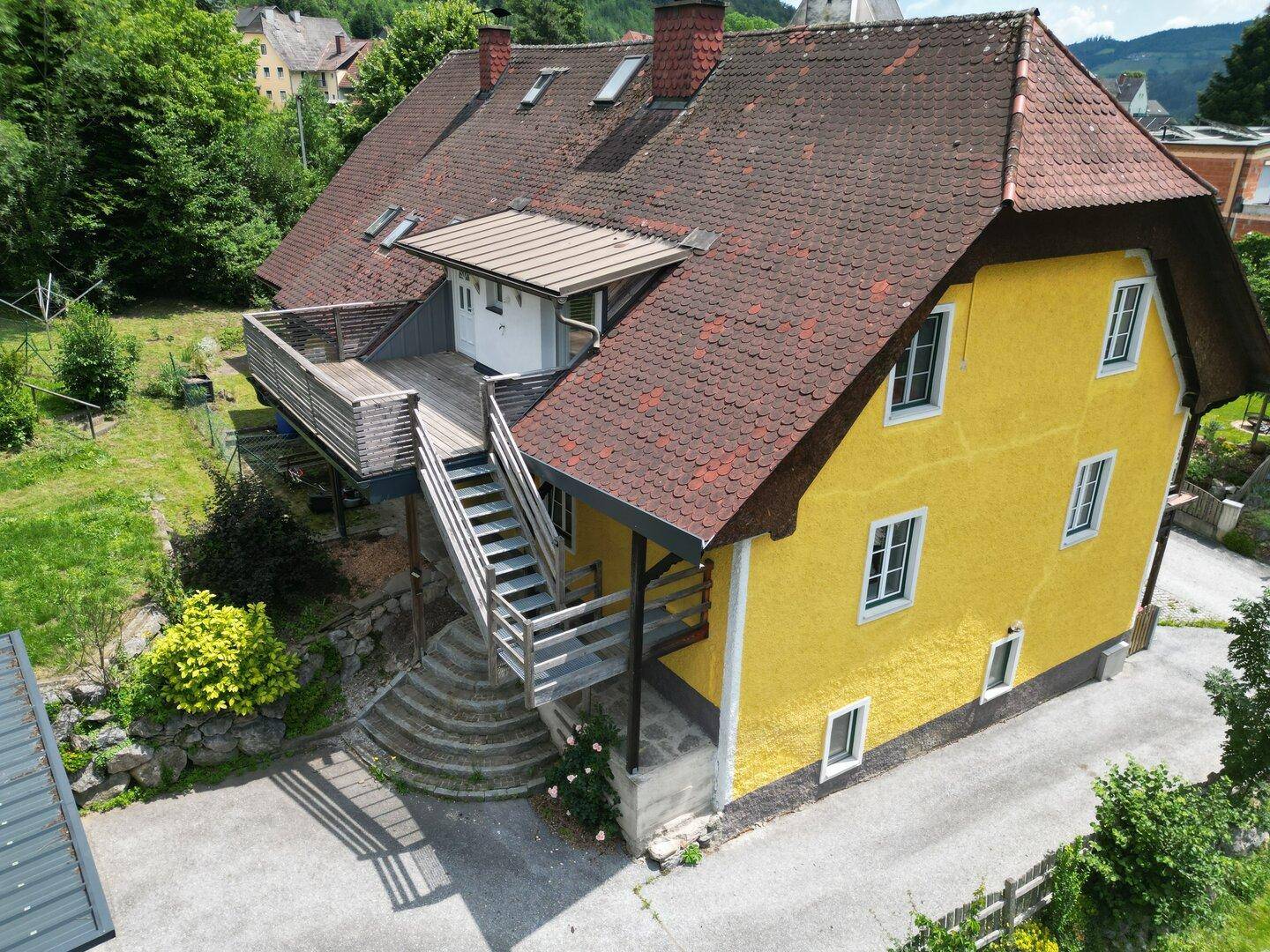Ansicht des gelben Hauses mit rotem Ziegeldach und einer Holzterrasse mit Außentreppe.