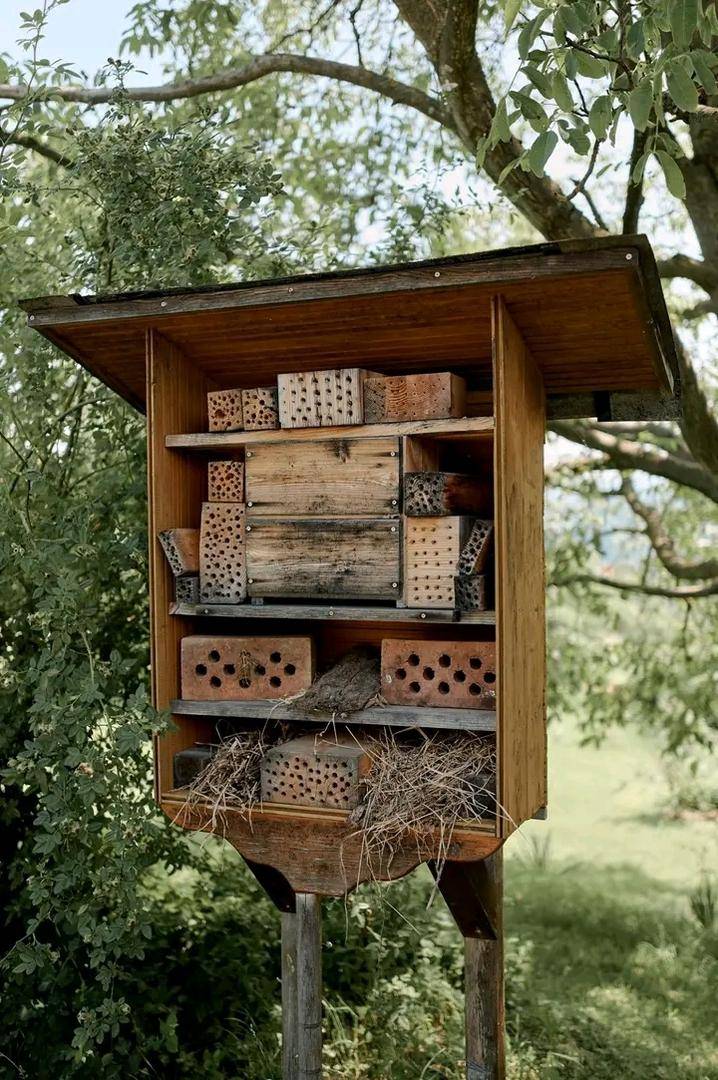 Ein großes Insektenhotel aus Holz und Ziegeln, umgeben von grünen Bäumen im Garten.