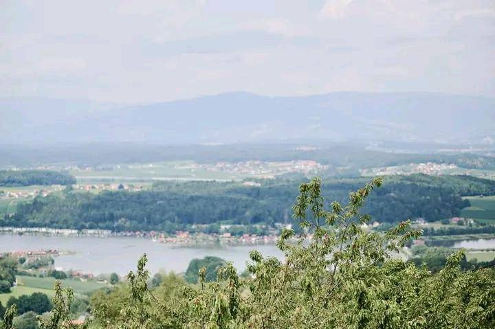 Weitläufige Landschaftsansicht mit einem See, umgeben von Wäldern und einem Dorf, sowie Bergen am Horizont.