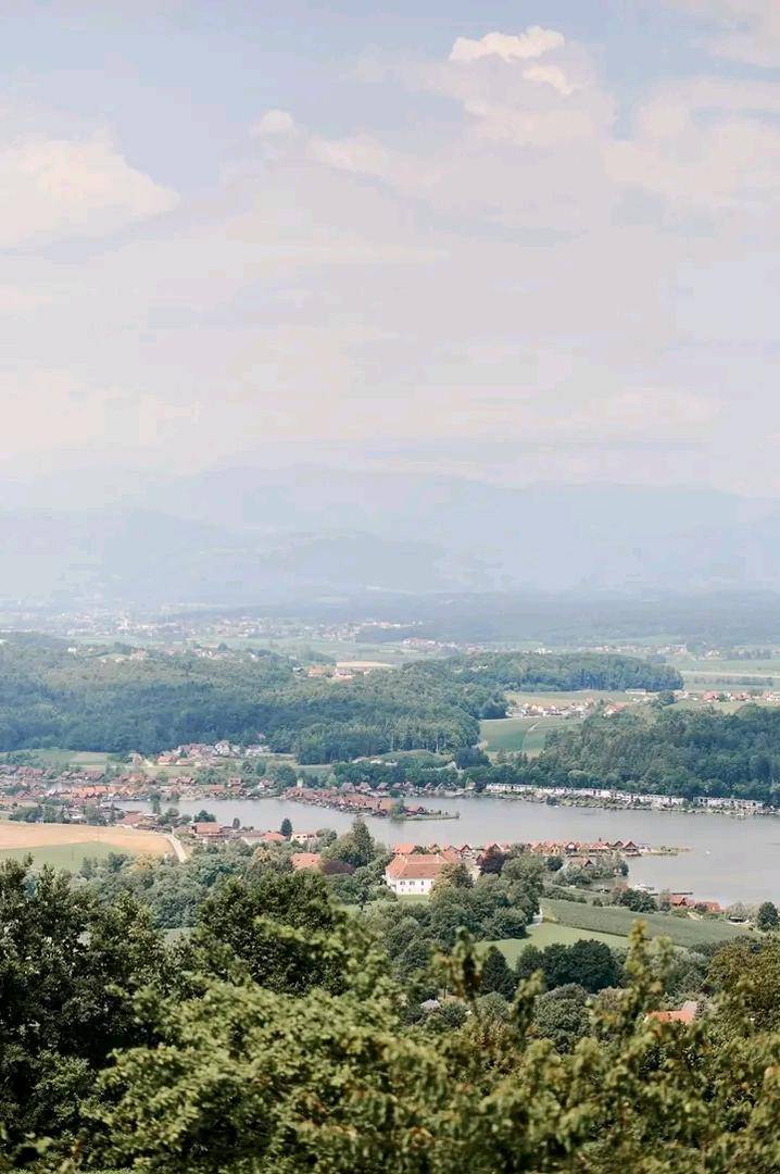 Panoramablick über eine weite Landschaft mit einem See, einem Dorf und sanften Hügeln im Hintergrund.