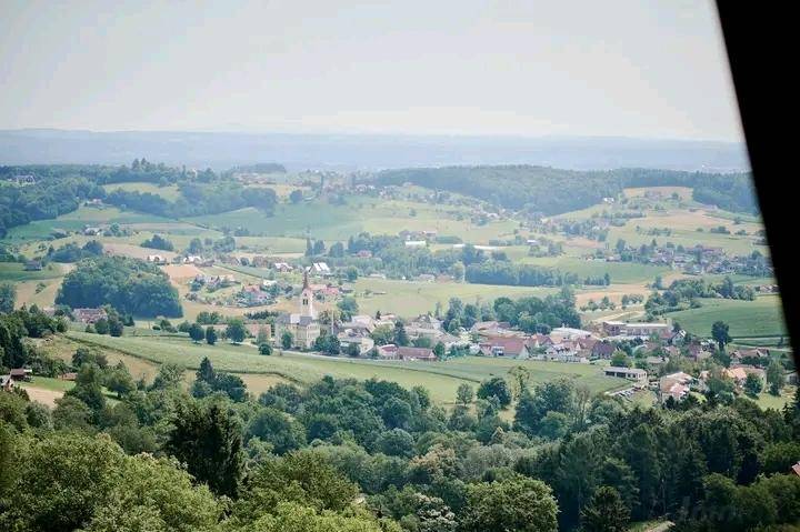Panoramablick auf ein malerisches Dorf, eingebettet in grüne Hügel und Wälder unter blauem Himmel.