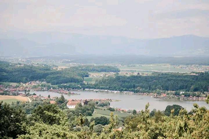 Breiter Panoramablick auf einen See, umgeben von grünen Wäldern und kleinen Siedlungen in der Ferne.