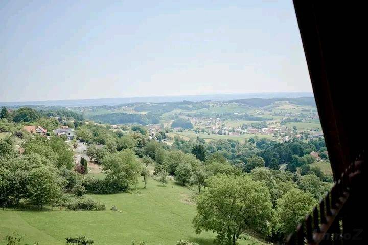 Weitläufige Landschaftsansicht mit grünen Hügeln, Wäldern und einem Dorf in der Ferne unter blauem Himmel.